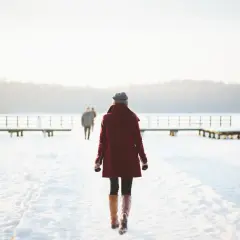 Kobieta w bordowym płaszczu idzie przez zaśnieżony teren, jakby uciekała od trudności, symbolizując nieumiejętność przyjmowania krytyki.