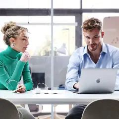 Kobieta w zielonym golfie słucha mężczyzny przy laptopie. Dyskutują, wymieniając konstruktywną krytykę, by wspólnie osiągnąć cel.