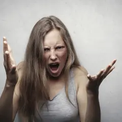 Młoda kobieta z rozczochranymi włosami krzyczy, jej twarz wykrzywiona w gniewie. Wygląda, jakby łatwo wpadała w gniew, gestykulując rękami.