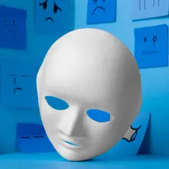 Biała maska na tle niebieskich karteczek z rysunkami smutnych buziek. Symbolizuje syndrom oszusta i jego przyczyny.