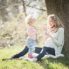 Ciężarna kobieta i małe dziecko puszczają bańki mydlane pod drzewem. Mama uśmiecha się, wiedząc, że robi wszystko, żeby cię chronić.