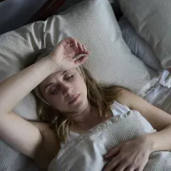 Kobieta leży w łóżku, czując się źle. Jej czoło jest wilgotne, co sugeruje nocne poty.