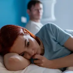 Kobieta z rudymi włosami leży smutna na poduszce, podczas gdy w tle widać niewyraźną postać mężczyzny. Czuć atmosferę toksycznego faceta i jego wpływu na nią.