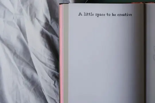 Otwarta książka z napisem "A little space to be creative" na białej stronie, obok pogniecionej szarej pościeli. Jak być kreatywnym?