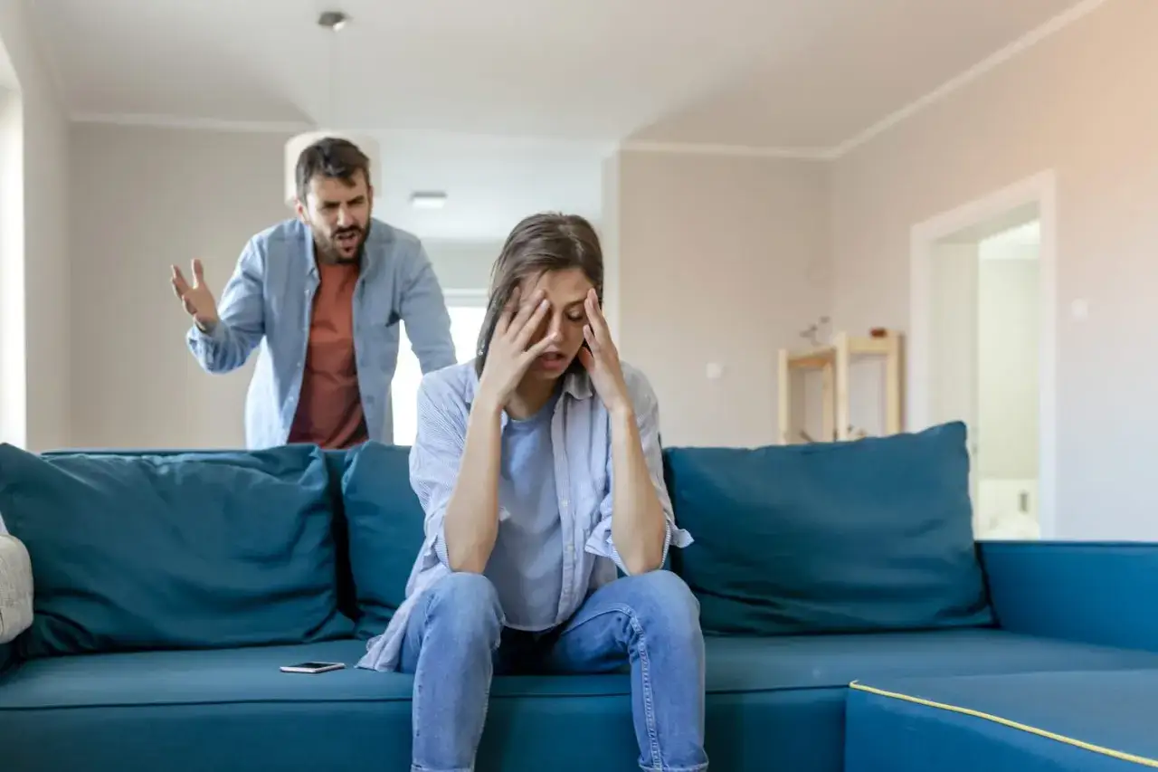 Kobieta z rękami na twarzy, przygnębiona, podczas gdy mężczyzna krzyczy za nią. Klasyczny przykład wypominania w psychologii relacji.